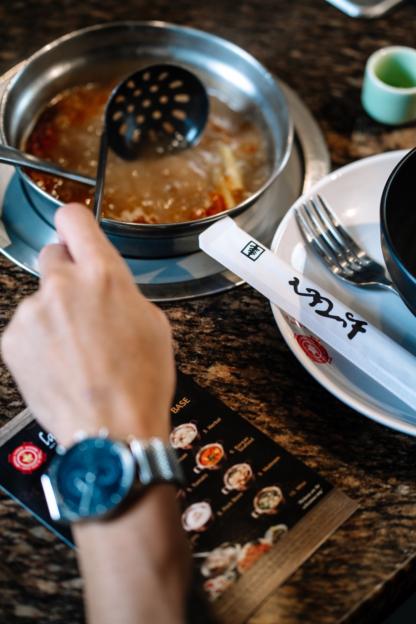 A ladle lifting from a simmering spicy hot pot broth beside Lanjie's soup-base menu card